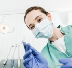The photo is taken from the point of view of a patient sitting in the dental chair. The registered dental hygienist (RDH), wearing blue gloves and light green scrubs, and light blue mask is holding up a dental instrument as they begin the procedure.