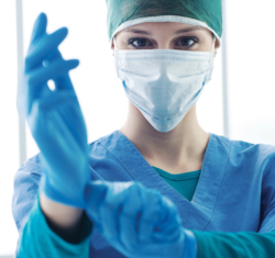 A woman donning personal protective equipment, a green cap, a light blue face mask, dark blue scrubs – pulling on a pair of dark blue exam gloves.