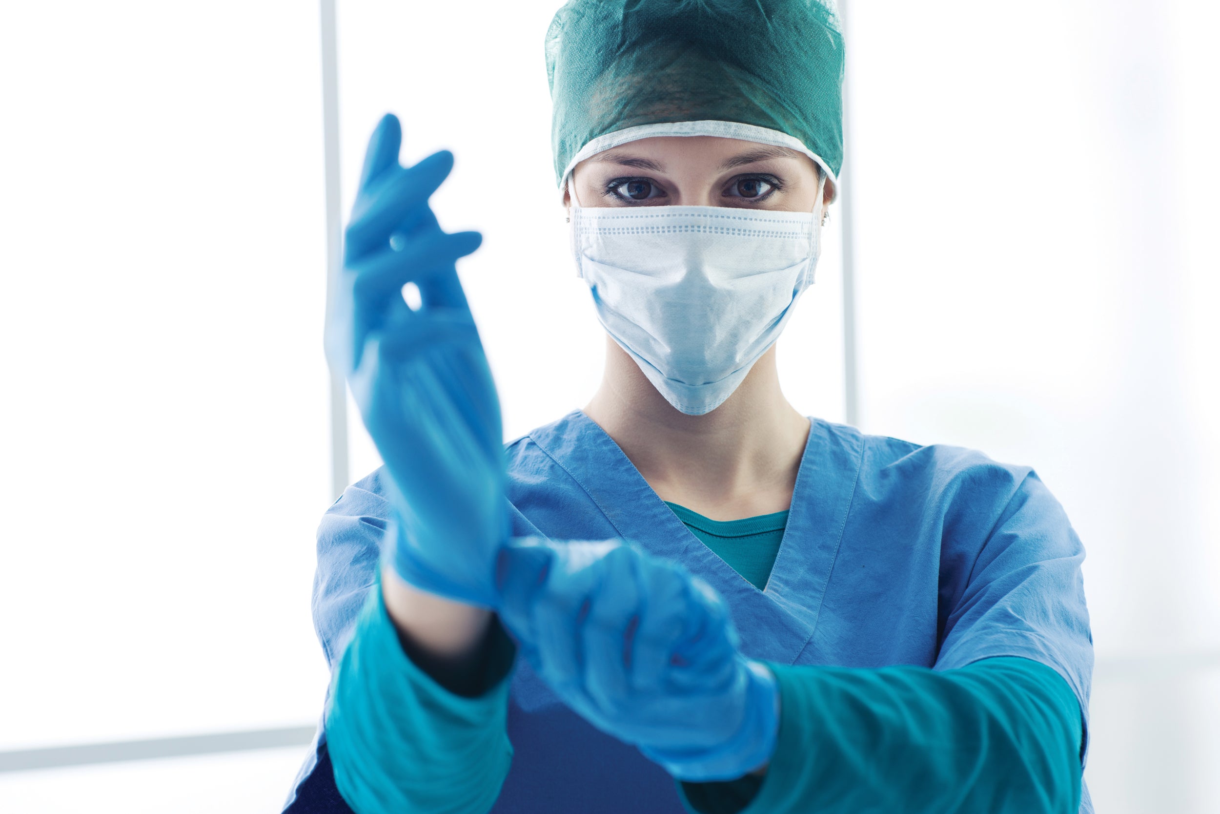 A woman donning personal protective equipment, a green cap, a light blue face mask, dark blue scrubs – pulling on a pair of dark blue exam gloves.