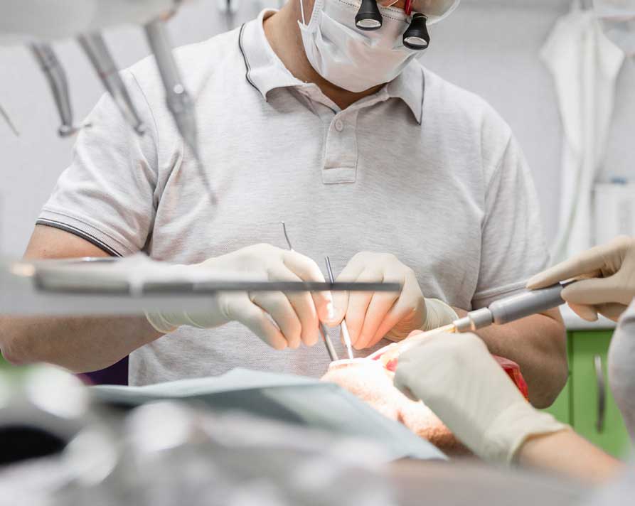 A dentist examining a patient's mouth is the focus of the image. We see only the hands of an assistant aiding the doctor. The dentist and assistant are both wearing natural colored NRL (natural rubber gloves).