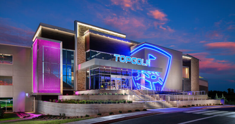 An exterior photo taken of a TopGolf venue at dusk. The blue and purple neon lights highlight the modern architecture of the building. The angles and lights add a lot of energy to the building, capturing the essence of the space.