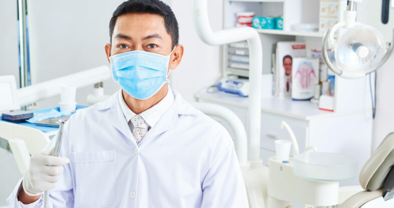 A dentist stands in a bright, light dental operatory holding a handpiece. In the very light colored background, the dentist, wearing a blue mask, carries the focal point of the image.