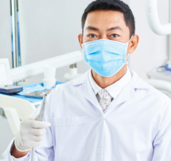 A dentist stands in a bright, light dental operatory holding a handpiece. In the very light colored background, the dentist, wearing a blue mask, carries the focal point of the image.