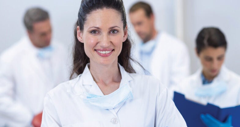 A woman doctor with dark hair smiles at the camera. She is wearing a white lab coat and has a mask around her neck. Behind her, out of focus, we can see three additional people wearing similar clothing referencing paperwork in front of them. They are in a bright white room.