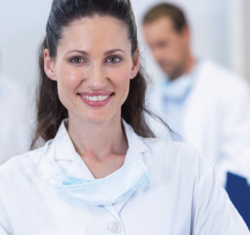 A woman doctor with dark hair smiles at the camera. She is wearing a white lab coat and has a mask around her neck. Behind her, out of focus, we can see three additional people wearing similar clothing referencing paperwork in front of them. They are in a bright white room.