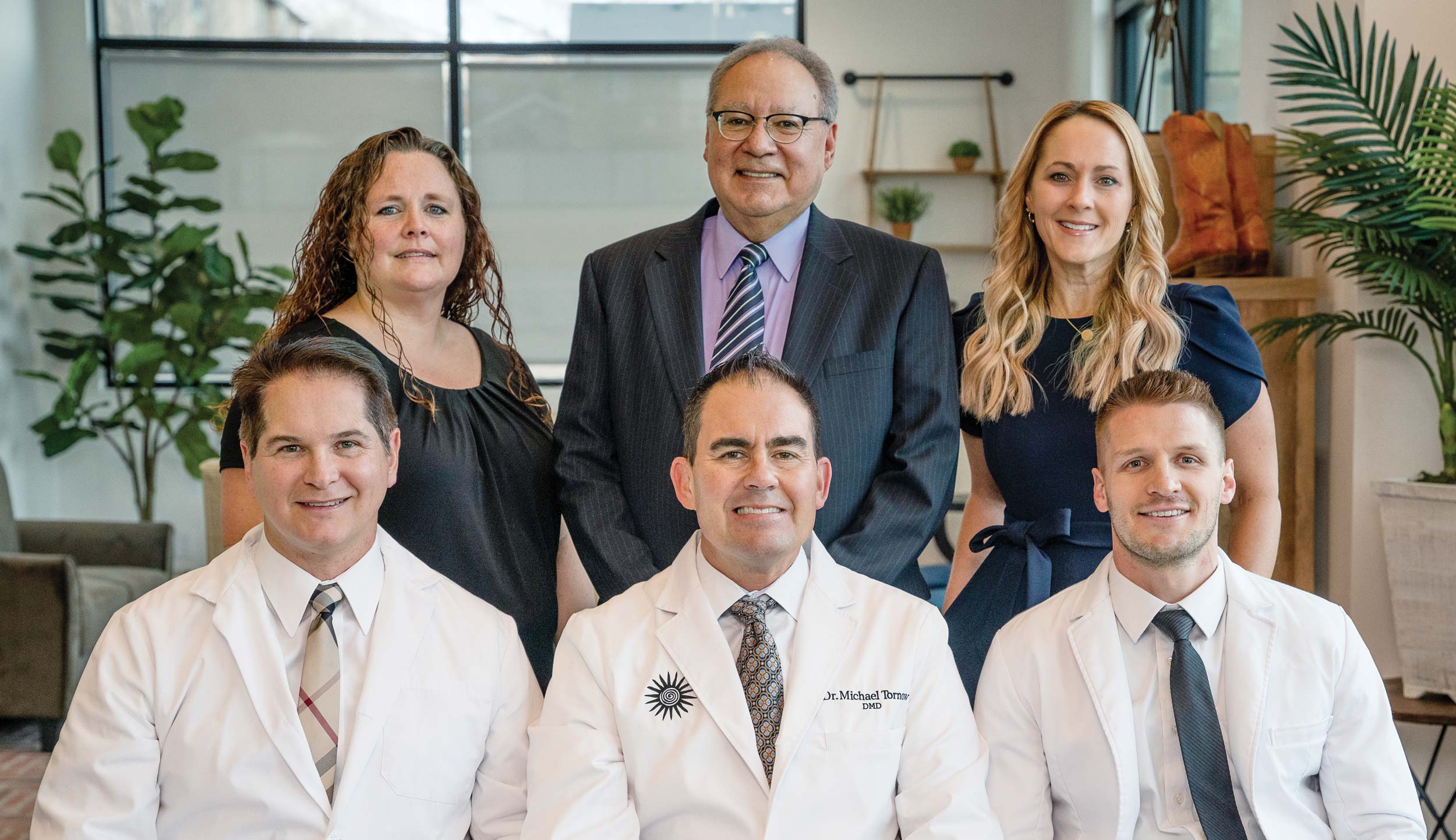 An interior group shot of the three managing partners and the director of finance and operations for Pony Express and the two integral Burkhart team members. The three doctors sit in a row smiling in their white coats. The second row of people are clustered behind them.