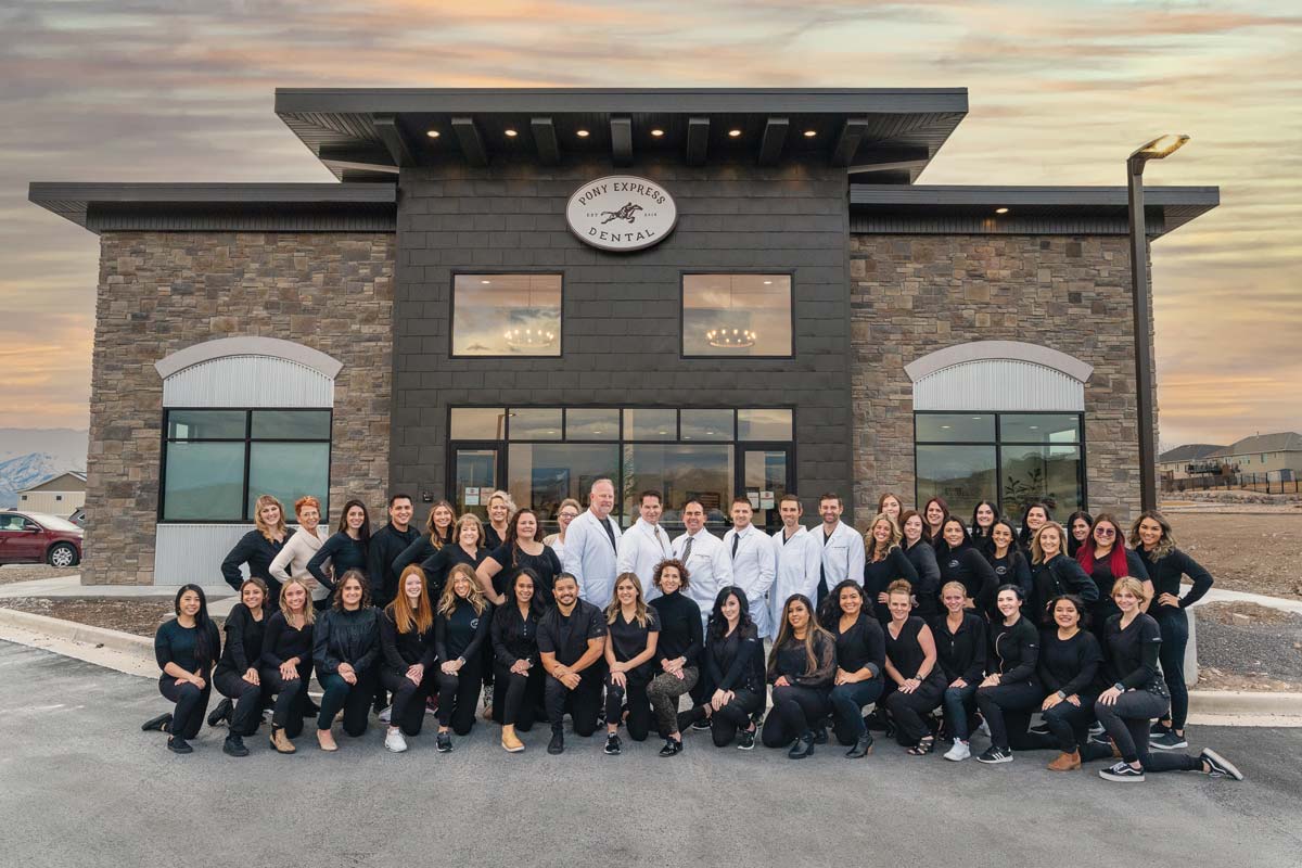 The Pony Express team – a group photo of 45 people. The group in the back is standing, the six doctors wearing white lab coats are at the heart of the back row. Their white coats bright against the dark, rustic exterior of the Pony Express dental practice. They are surrounded by the rest of the team dressed in black. 
