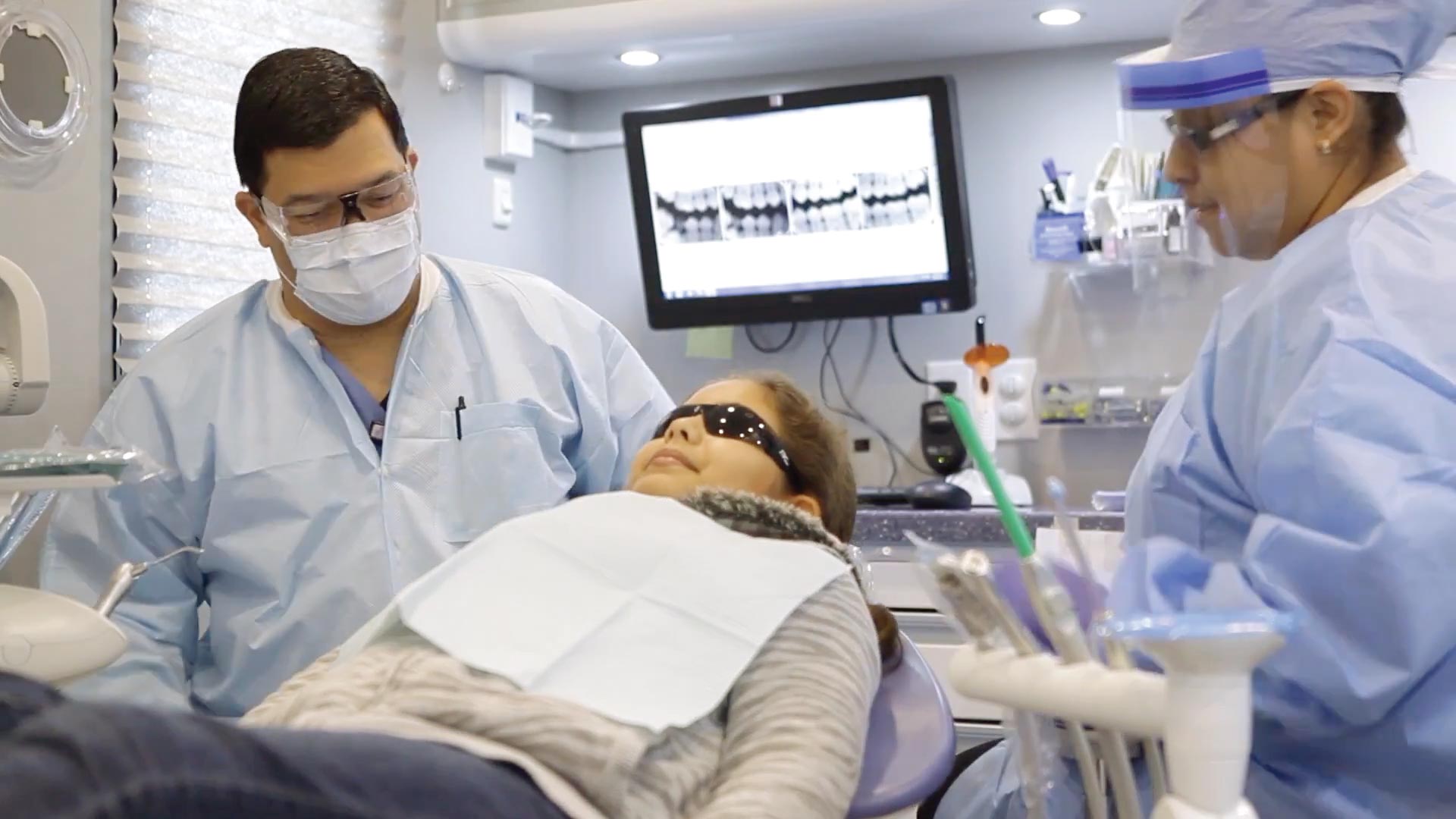 Team members at St. David's Foundation prep an elementary school age girl for dental care. The patient is reclined in an operatory chair, she wears wrap-around sunglasses to protect her eyes from the light. The doctor sits over the right shoulder of the girl. He's wearing safety glasses, a mask, and a scrubs. Over the girl's left shoulder is an assistant wearing a face shield, glasses, a blue hair cover and blue scrubs. On a tv screen behind the patient are four x-ray images.