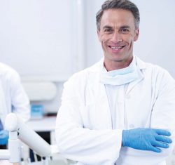 Male dentist smiles broadly at the camera. Around his neck he's wearing a mask, dressed in a white doctor's coat with blue gloves, he stands in the foreground of the image. Out of focus in the background a team member addresses the needs of a patient reclined in a dental chair. The whole space is bright and light.