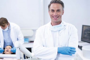 Male dentist smiles broadly at the camera. Around his neck he's wearing a mask, dressed in a white doctor's coat with blue gloves, he stands in the foreground of the image. Out of focus in the background a team member addresses the needs of a patient reclined in a dental chair. The whole space is bright and light.