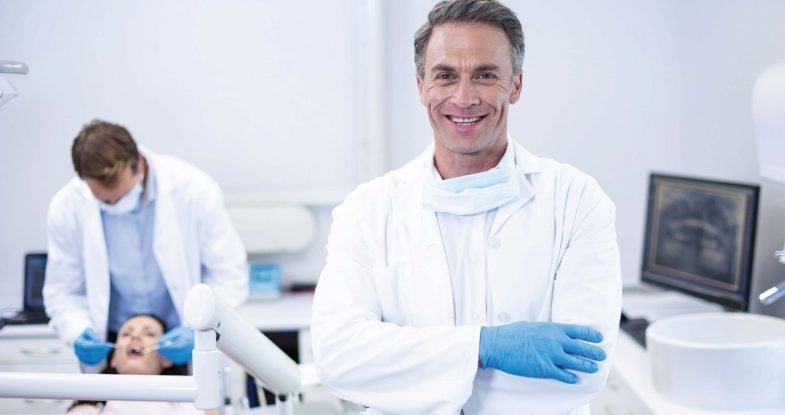 Male dentist smiles broadly at the camera. Around his neck he's wearing a mask, dressed in a white doctor's coat with blue gloves, he stands in the foreground of the image. Out of focus in the background a team member addresses the needs of a patient reclined in a dental chair. The whole space is bright and light.