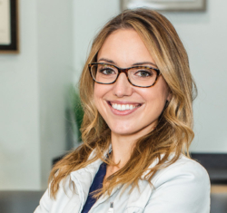 Dr. Samantha Mize of Bitesize Dentistry. She is standing in a quarter turn, smiling toward the camera wearing her white doctor's coat. Her blond hair is down, nicely styled for an approachable look. She is wearing glasses and standing in front of the practice's reception desk.