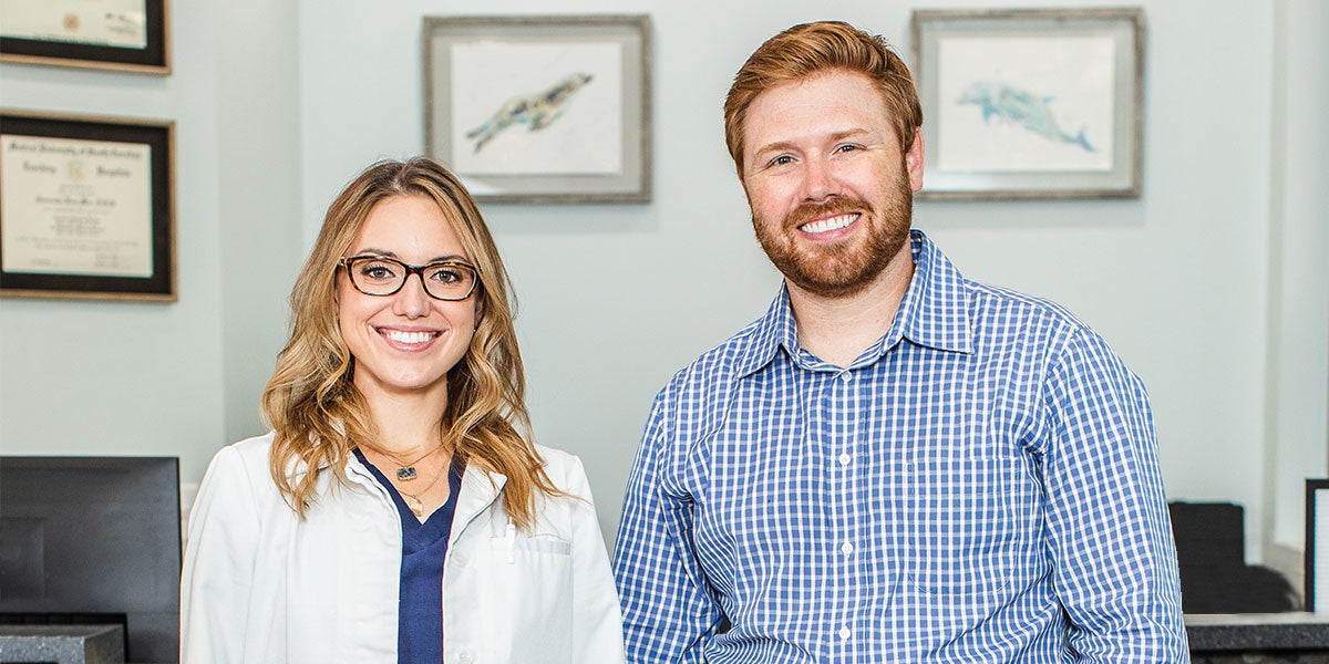 Dr. Samantha Mize and her husband Josh both work at Dr. Sam’s practice called Bitesize Pediatric Dentistry in Anchorage, Alaska. Pictured here, they both stand shoulder to shoulder smiling at the camera in front of their reception desk at the practice.