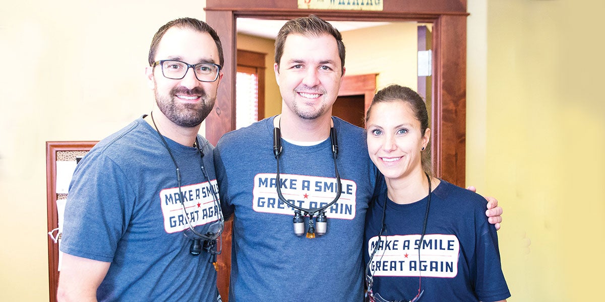 From left to right: Dr. Cody Winterholler, Dr. William Winterholler (Dr. Will), and staff member. The trio stands close together with arms around each other smiling at the camera. They are all wearing "Make a Smile Great Again" t-shirts in honor of their event. 