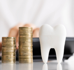 Finances are one of the main reasons patients delay treatment. This image focuses on that one reason. We see the hands of a woman calculating numbers behind stair step stacks of coins. Next to the last stack is a toy tooth standing slightly higher than the stack of coins illustrating how dental treatment can be just out of reach for some.